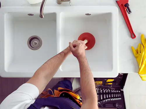 Plumber using plunger to clear clogged kitchen sink drain with tools and gloves nearby